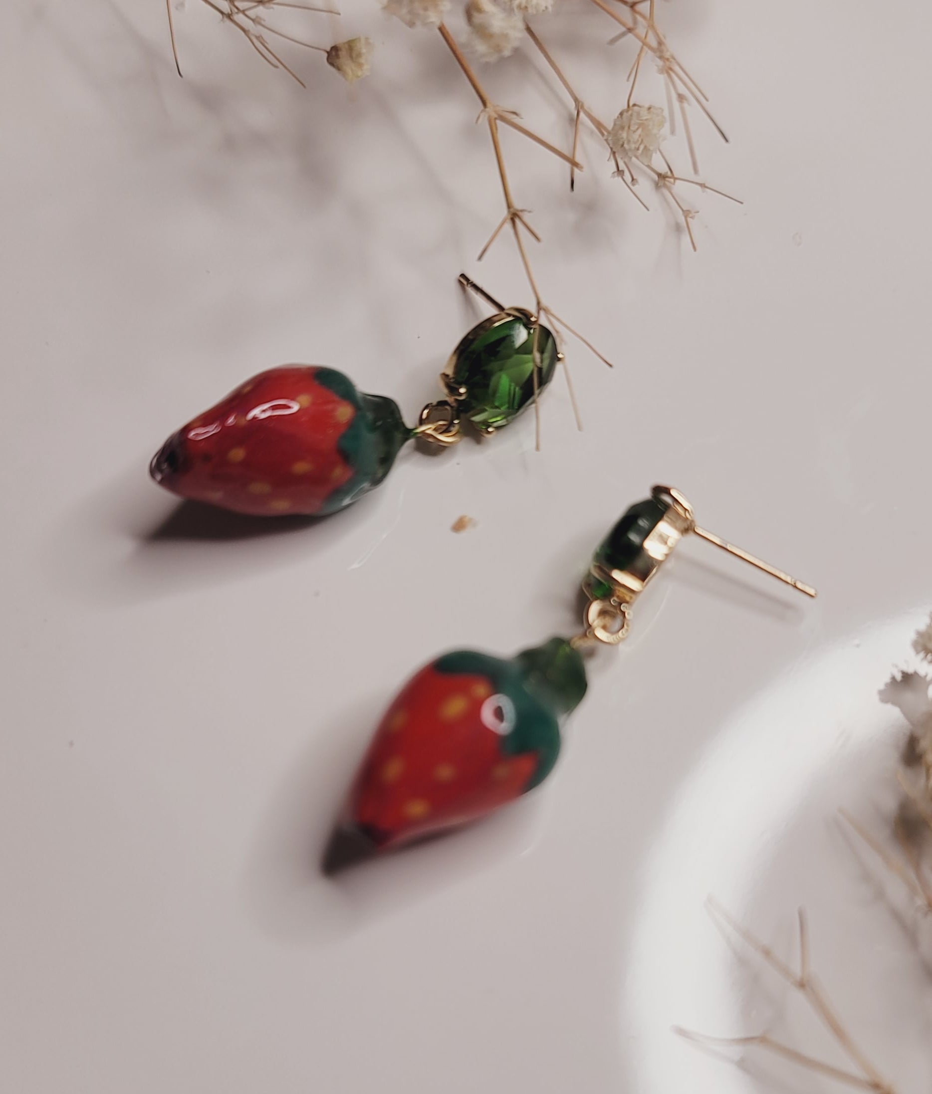 Pair of strawberry-shaped earrings on a light surface with dried flowers.
