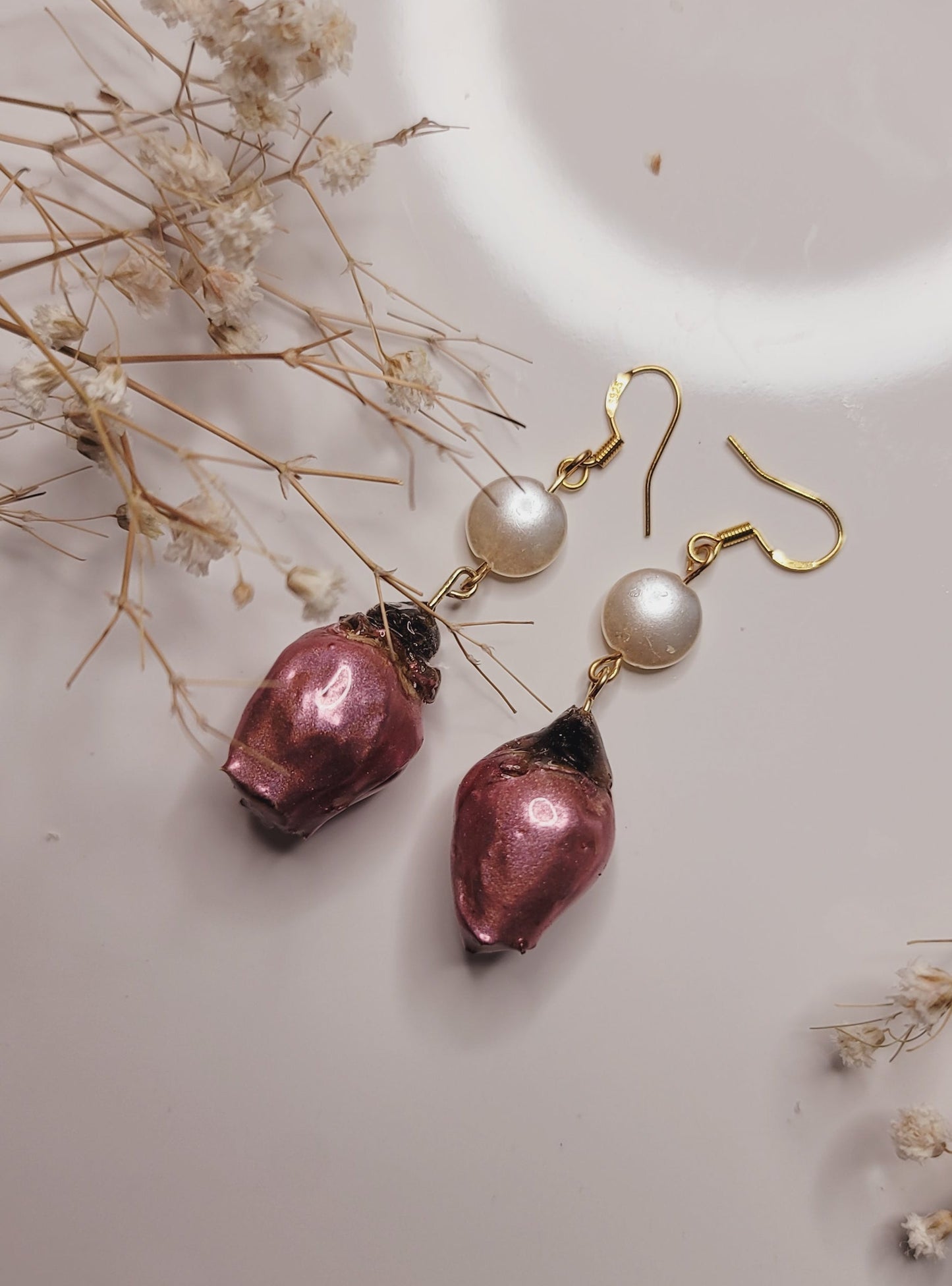Pair of earrings with pink and pearl-like stones on a light surface with dried flowers.
