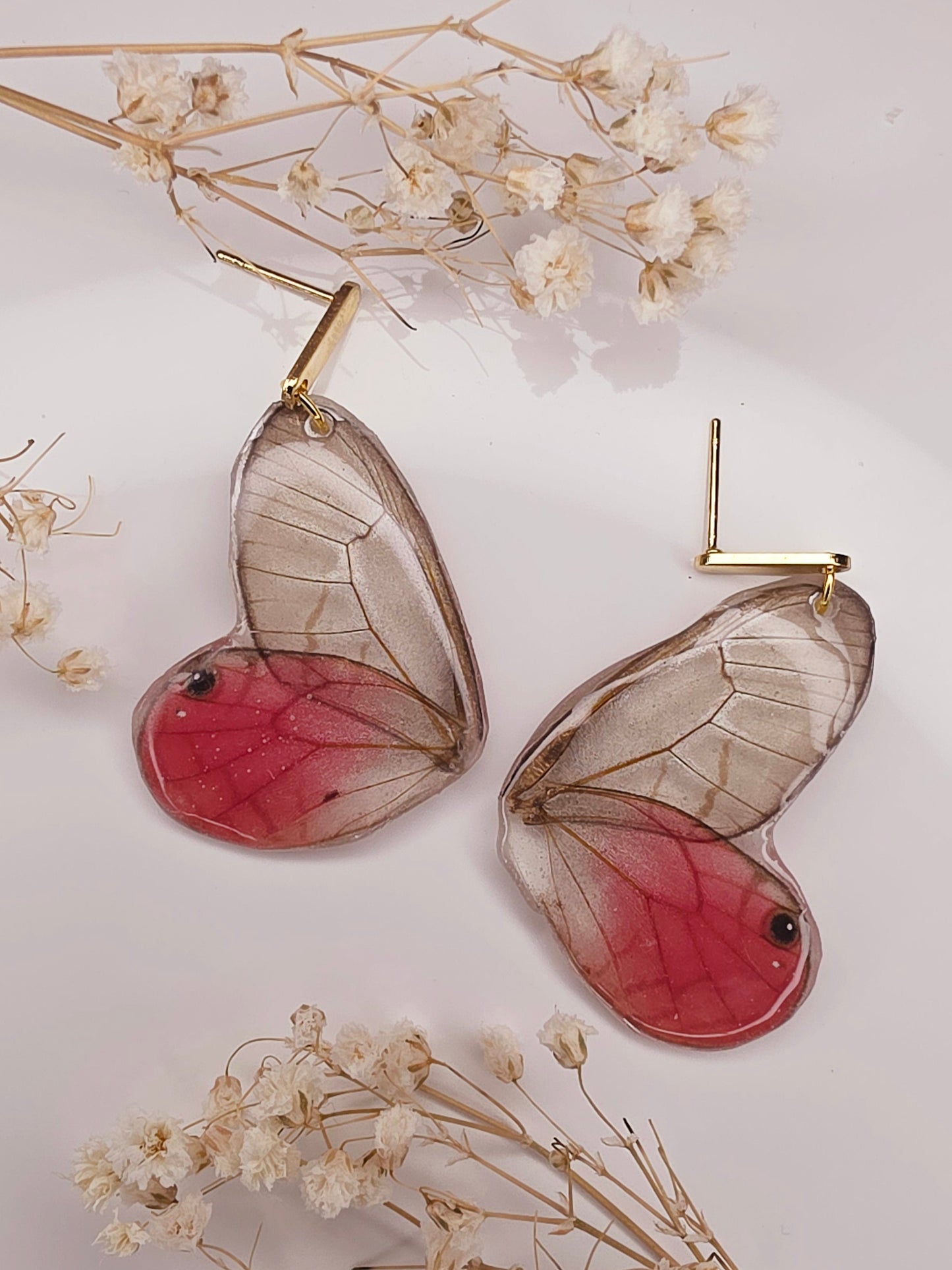 Butterfly-shaped earrings with red centers and clear wings on a light background with dried flowers.