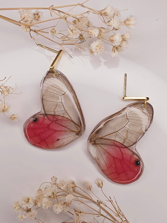 Butterfly-shaped earrings with red centers and clear wings on a light background with dried flowers.