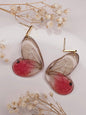 Butterfly-shaped earrings with red centers and clear wings on a light background with dried flowers.